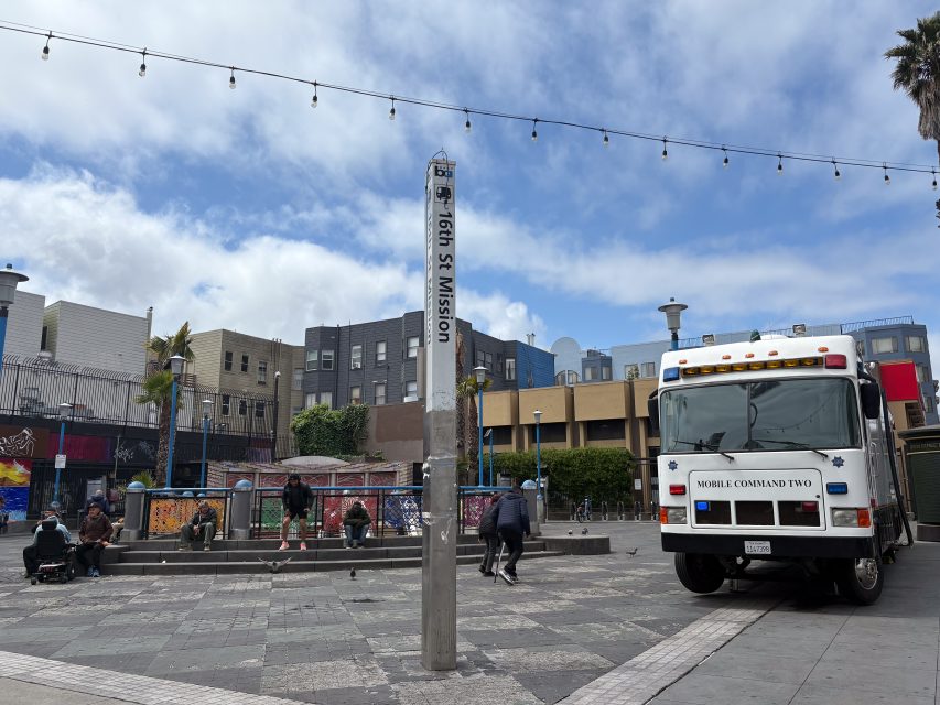 A city square with a pole labeled "16th St Mission," people sitting and walking, and a vehicle marked "Mobile Command Two" parked nearby.