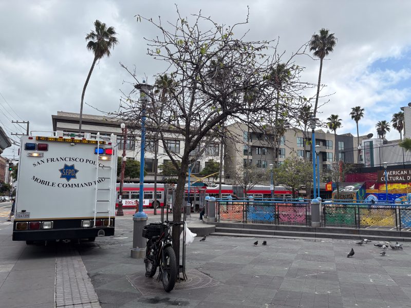A San Francisco police mobile command vehicle is parked near a tree and a bike in an urban plaza with pigeons and buildings in the background.