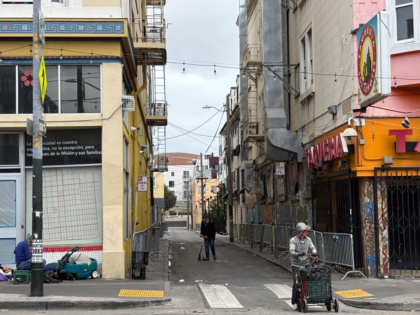 A narrow urban alleyway with a person sweeping in the distance, another person with a walker crossing the street, and various buildings on both sides.