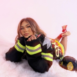 A woman lies on a white fluffy surface, wearing a firefighter jacket and red high heels. A firefighter helmet is placed nearby.