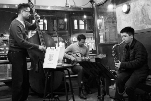Three musicians perform indoors; one plays upright bass, another plays guitar, and the third plays saxophone. The scene is in black and white.