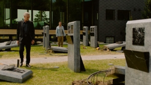 Two men stand among several damaged and toppled gravestones on a patch of grass near a modern building with large windows.