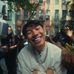 A person wearing a white shirt and chain necklace smiles with arms crossed, standing outdoors in front of a group and residential buildings.
