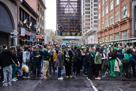 A large crowd of people in festive attire gather on a city street celebrating St. Patrick's Day. Buildings and street signs are visible in the background.