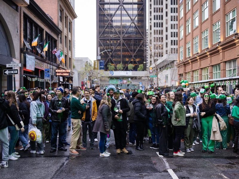 A large crowd of people in festive attire gather on a city street celebrating St. Patrick's Day. Buildings and street signs are visible in the background.