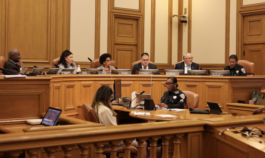 A group of officials sit at a raised dais in a wood-paneled room, while a uniformed officer and others sit below with laptops and documents.