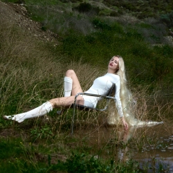 A person in a white outfit and boots relaxes on a chair in a grassy, wet area with a stream nearby.