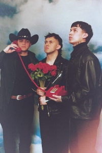 Three people in dark clothing stand against a cloudy backdrop. One holds a red phone and wears a cowboy hat, another holds roses, and the third gazes upwards.