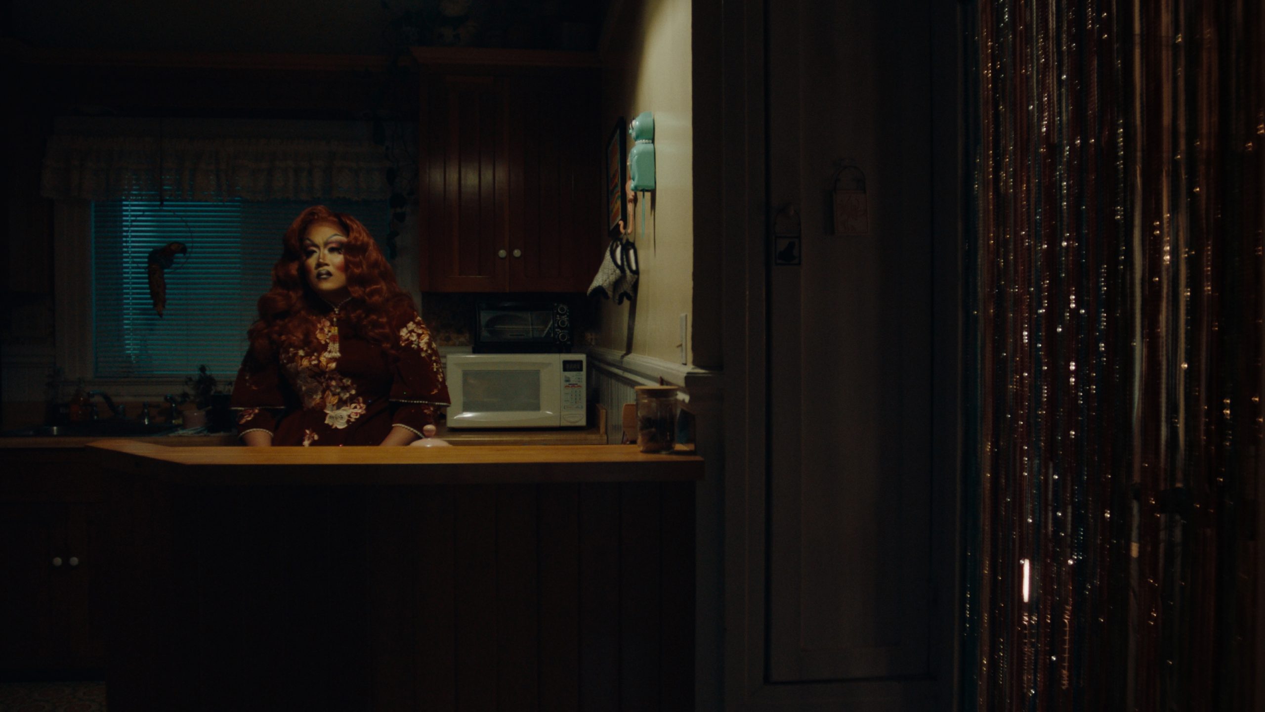 A person with red hair and makeup sits alone at a kitchen counter, illuminated by soft light, with a microwave and cabinets visible in the background.