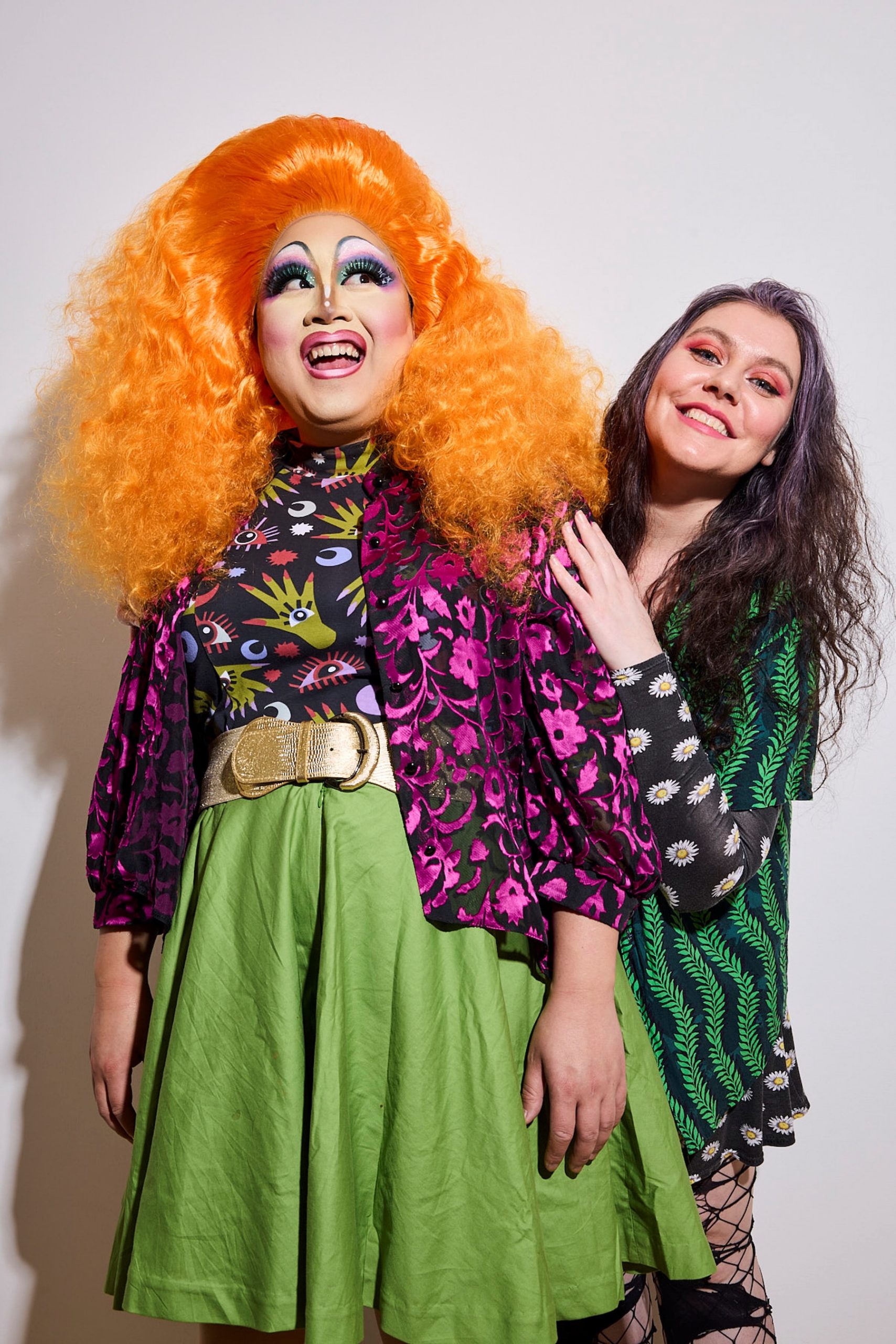 Two people stand together smiling. The person on the left wears a bright orange wig, colorful makeup, and vibrant clothes, while the person on the right has long, wavy hair and a floral dress.