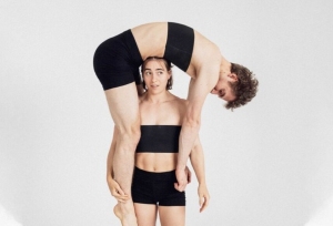 Two people in black athletic wear; one stands upright while the other is bent over their shoulders, creating a human arch.