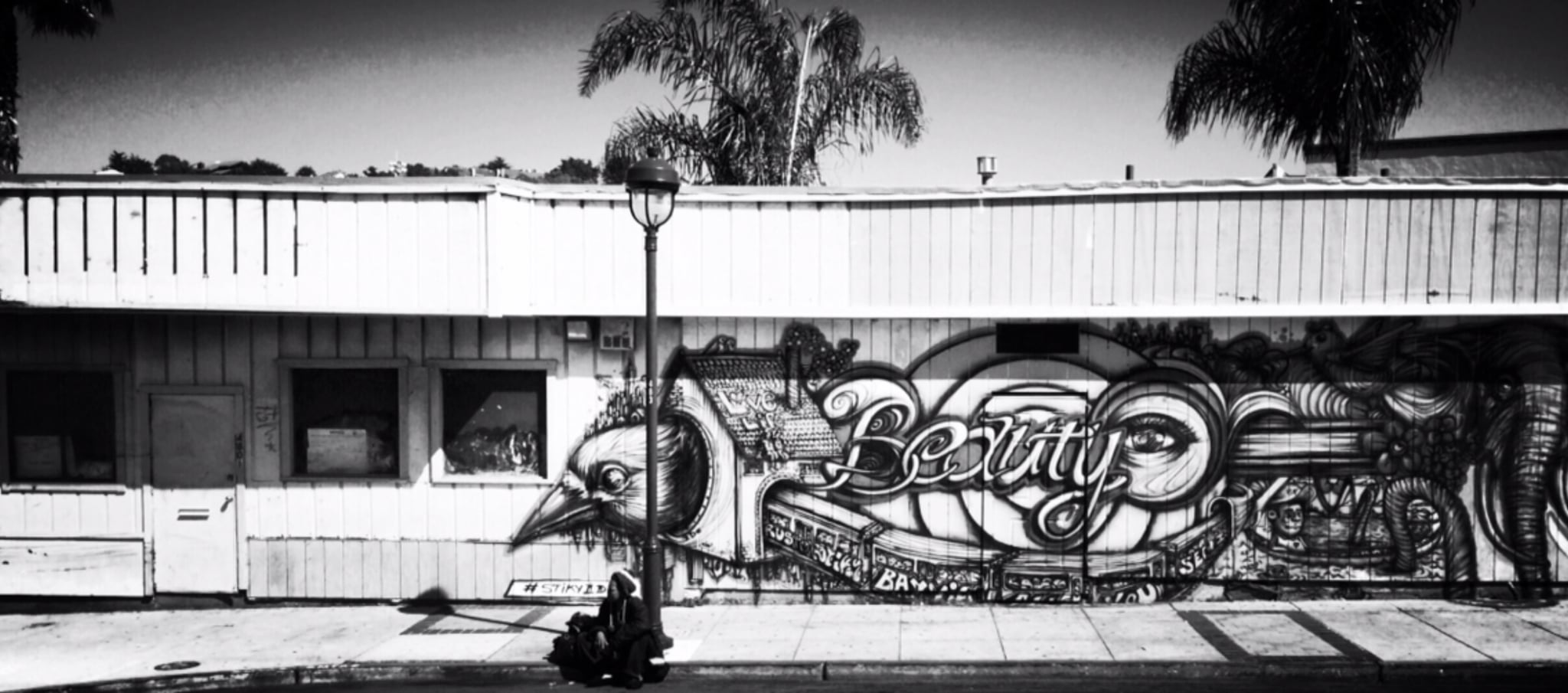 Black and white photo of a building with a mural featuring a bird and the word "beauty" painted on the exterior wall; palm trees and a streetlamp are visible.