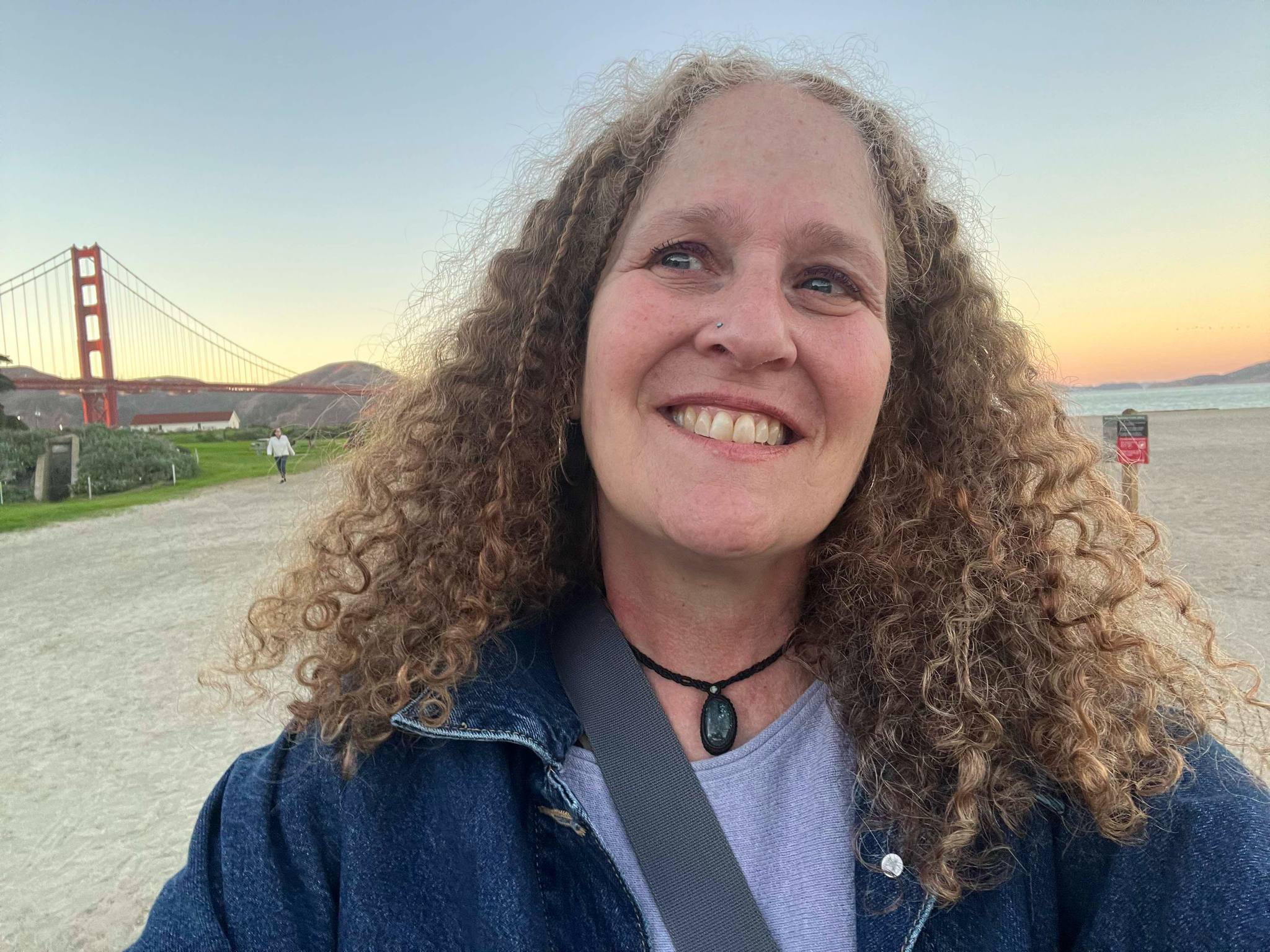 A person with curly hair smiles while taking a selfie at a beach with the Golden Gate Bridge visible in the background at sunset.