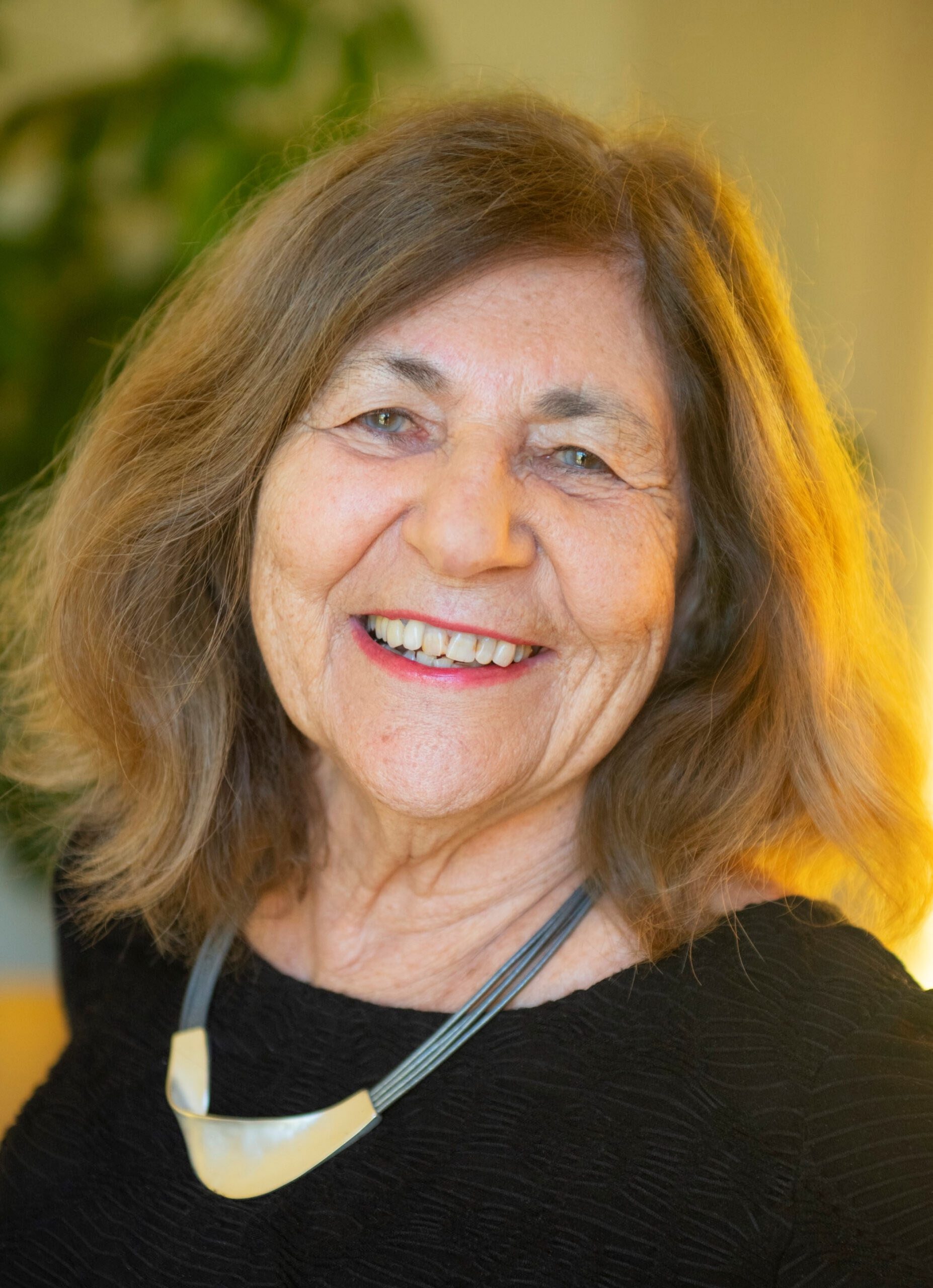 An older woman with shoulder-length brown hair and a black top smiles at the camera, wearing a modern silver necklace; blurred greenery is visible in the background.