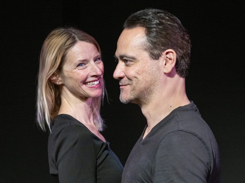 A woman and a man stand close together, facing each other and smiling. Both are dressed in black tops against a dark background.