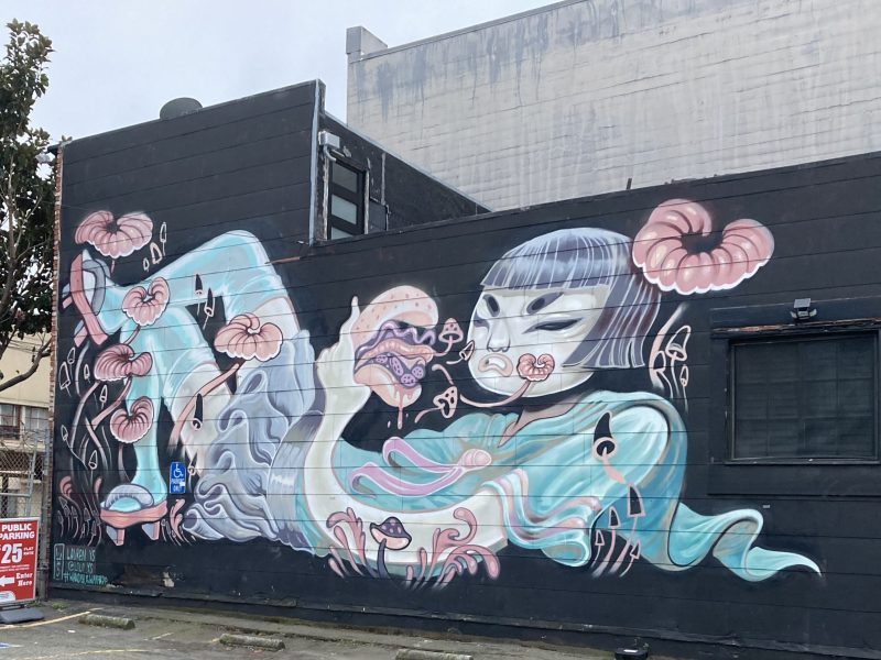 Mural on a dark building wall depicting a stylized figure with short hair, surrounded by large flowers, swirling teal shapes, and mushroom-like elements.