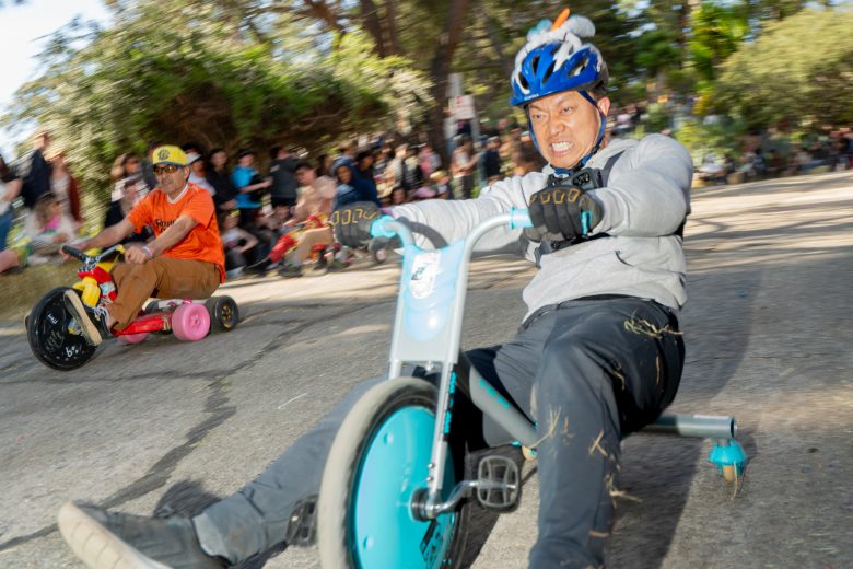 Two adults race on oversized tricycles down a sloped street, with one wearing a blue helmet in the foreground and a crowd of spectators in the background.