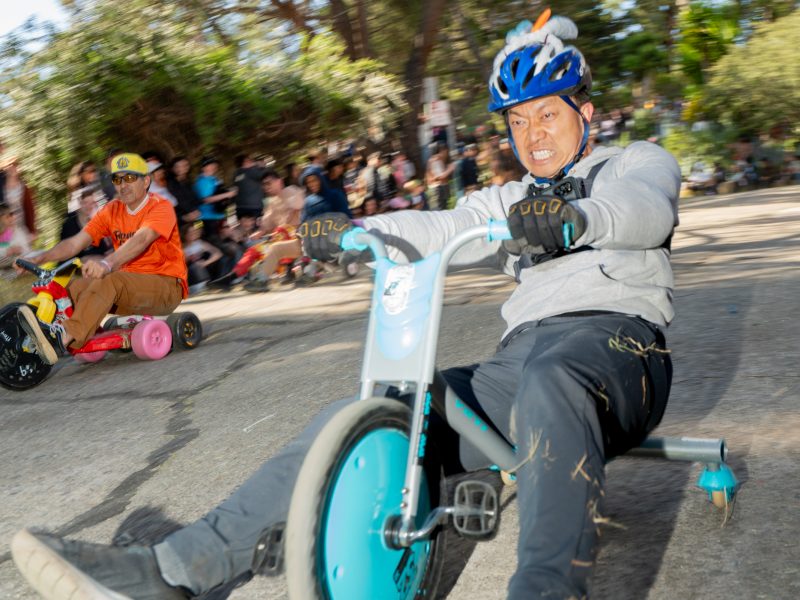 Two adults race on oversized tricycles down a sloped street, with one wearing a blue helmet in the foreground and a crowd of spectators in the background.