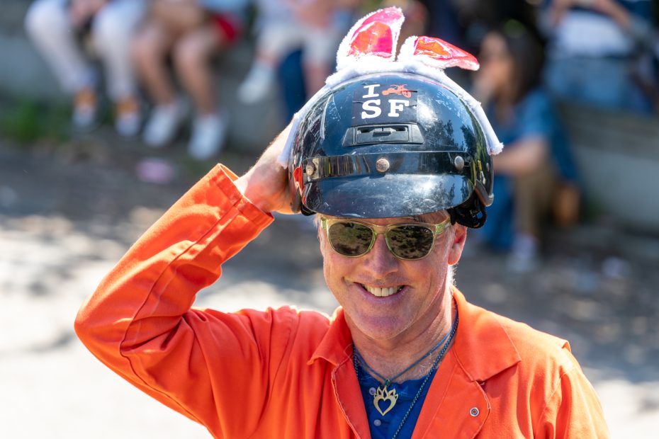 A man in an orange jumpsuit smiles, wearing sunglasses and a helmet reading "I SF" with bunny ears attached. People sit blurred in the background.