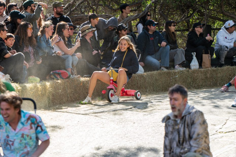 A woman rides a red tricycle down a paved path while a crowd of people sits on straw bales and watches outdoors.
