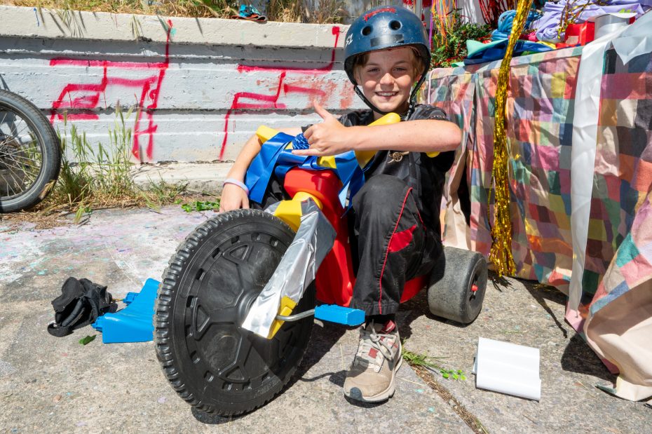 A child wearing a blue helmet sits on a homemade go-kart with a duct-taped front wheel, smiling and giving a thumbs-up.