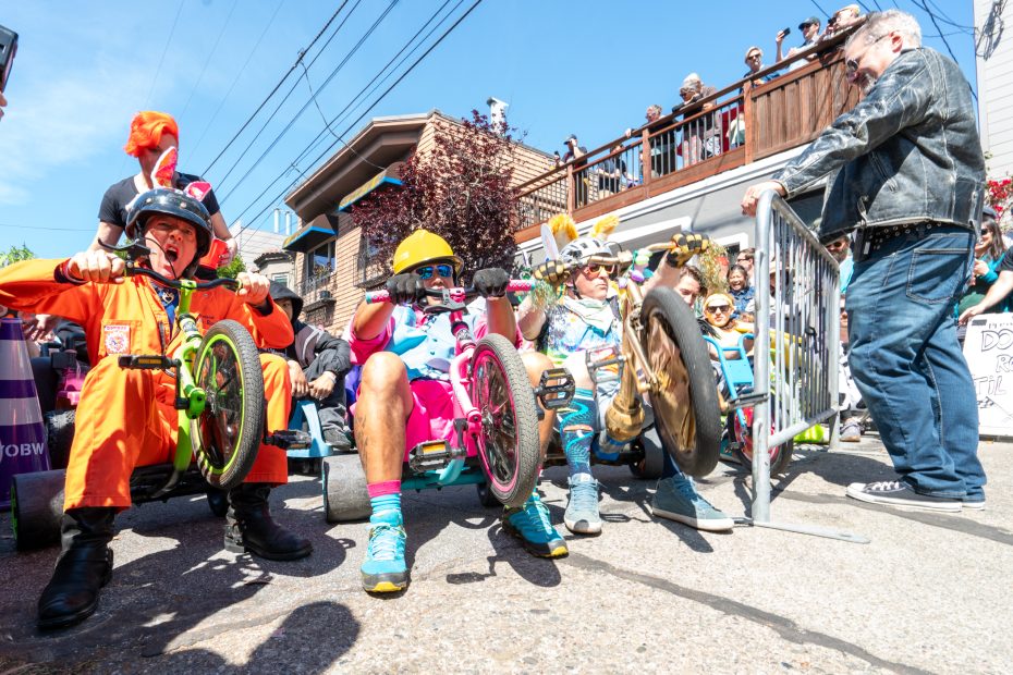 Adults in colorful costumes ride tricycles aggressively at a lively outdoor race, with spectators watching and a man holding a barrier nearby.