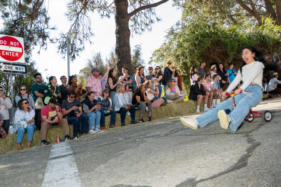A woman rides a small cart down a hill, smiling, as a crowd of people watches and takes photos; a “Do Not Enter” and “One Way” sign are visible in the background.