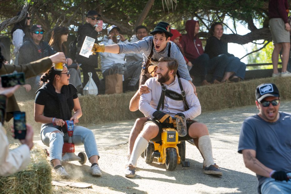 Two adults dressed in playful costumes ride a small toy vehicle down a hill, surrounded by cheering spectators and hay bales.