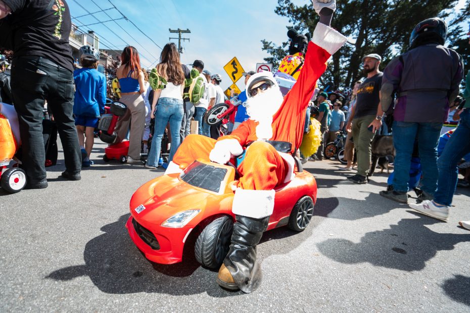 A person dressed as Santa Claus sits in a small red toy car, raising one arm, surrounded by a crowd outdoors on a sunny day.