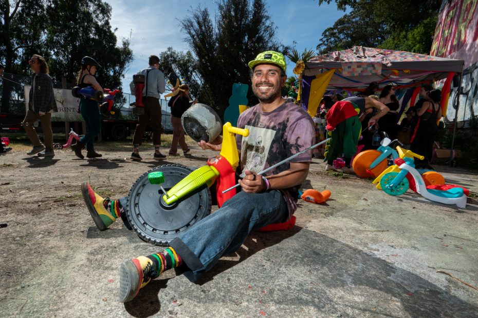 A smiling man in colorful clothes sits on a toy tricycle holding improvised objects, with people and play equipment in the background at an outdoor event.