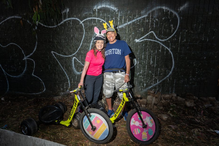 Two people wearing helmets and animal ear headbands stand with decorated yellow trikes in front of a graffiti-covered wall at night.