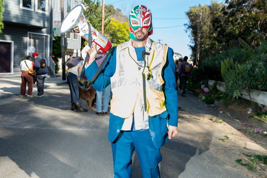 A man in a colorful luchador mask and reflective vest holds a megaphone while standing on a sunny street; people and trees are visible in the background.