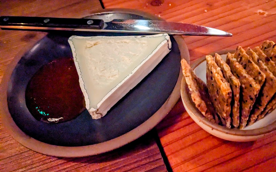 A wedge of cheese with a knife and fruit preserve on a plate, accompanied by a small bowl of sliced crackers, on a wooden table.