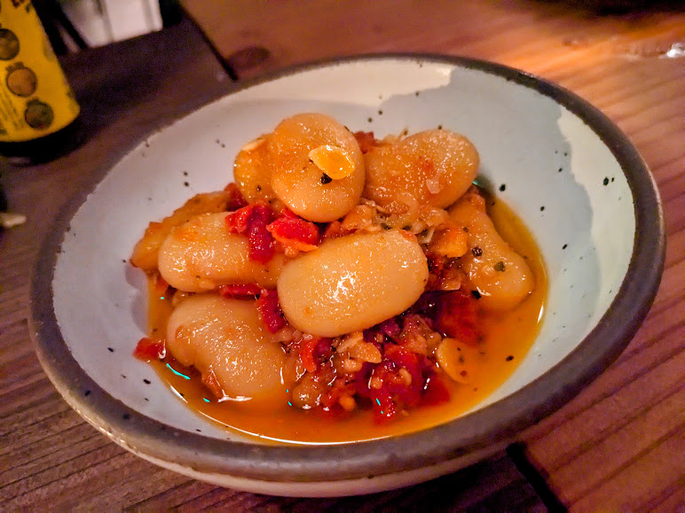 A bowl of large white beans in a tomato-based sauce, garnished with chopped vegetables, served in a ceramic dish on a wooden table.