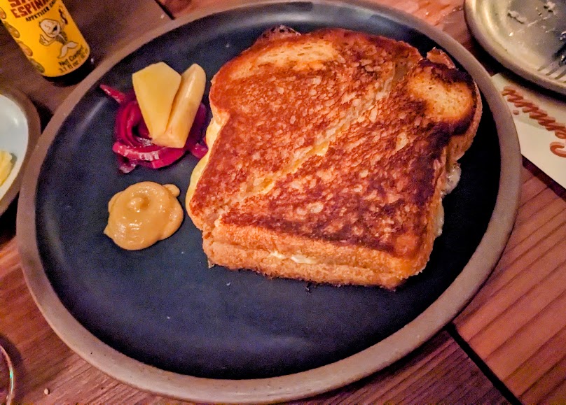 Grilled cheese sandwich on a dark plate with mustard, pickled vegetables, and jalapeños on the side.