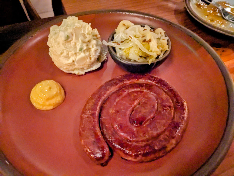 A coiled sausage with mustard, potato salad, and cabbage on a brown plate.