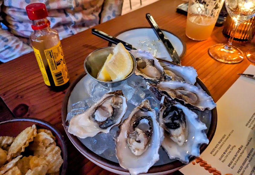 A plate of six oysters on ice with lemon wedges. A bottle of hot sauce, a glass of beer, and a menu are on the table.
