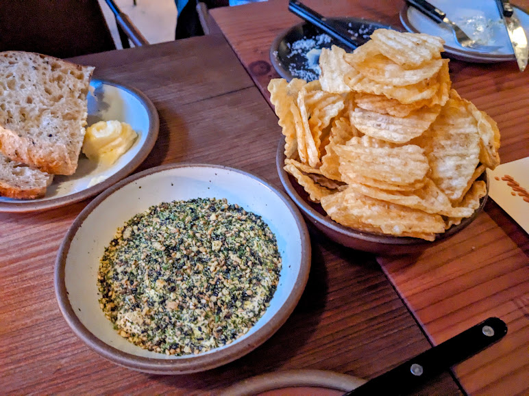 Plates of bread with butter, crinkle-cut potato chips, and a bowl of seasoning on a wooden table.