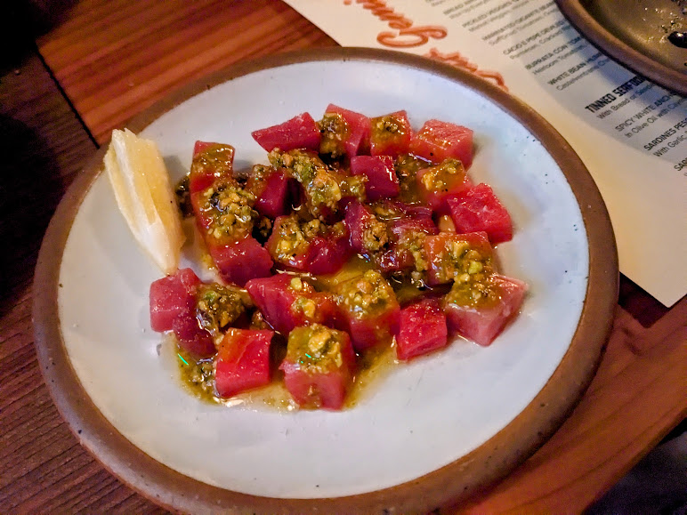 A plate of diced raw fish, likely tuna, topped with a sauce, is served alongside a wedge of lemon on a ceramic dish. A menu is visible in the background on a wooden table.