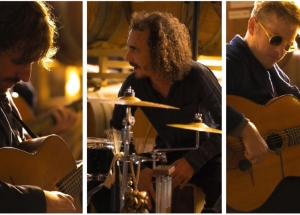 Three musicians perform indoors; two play guitars and one plays drums. The scene is warmly lit, creating a cozy atmosphere.