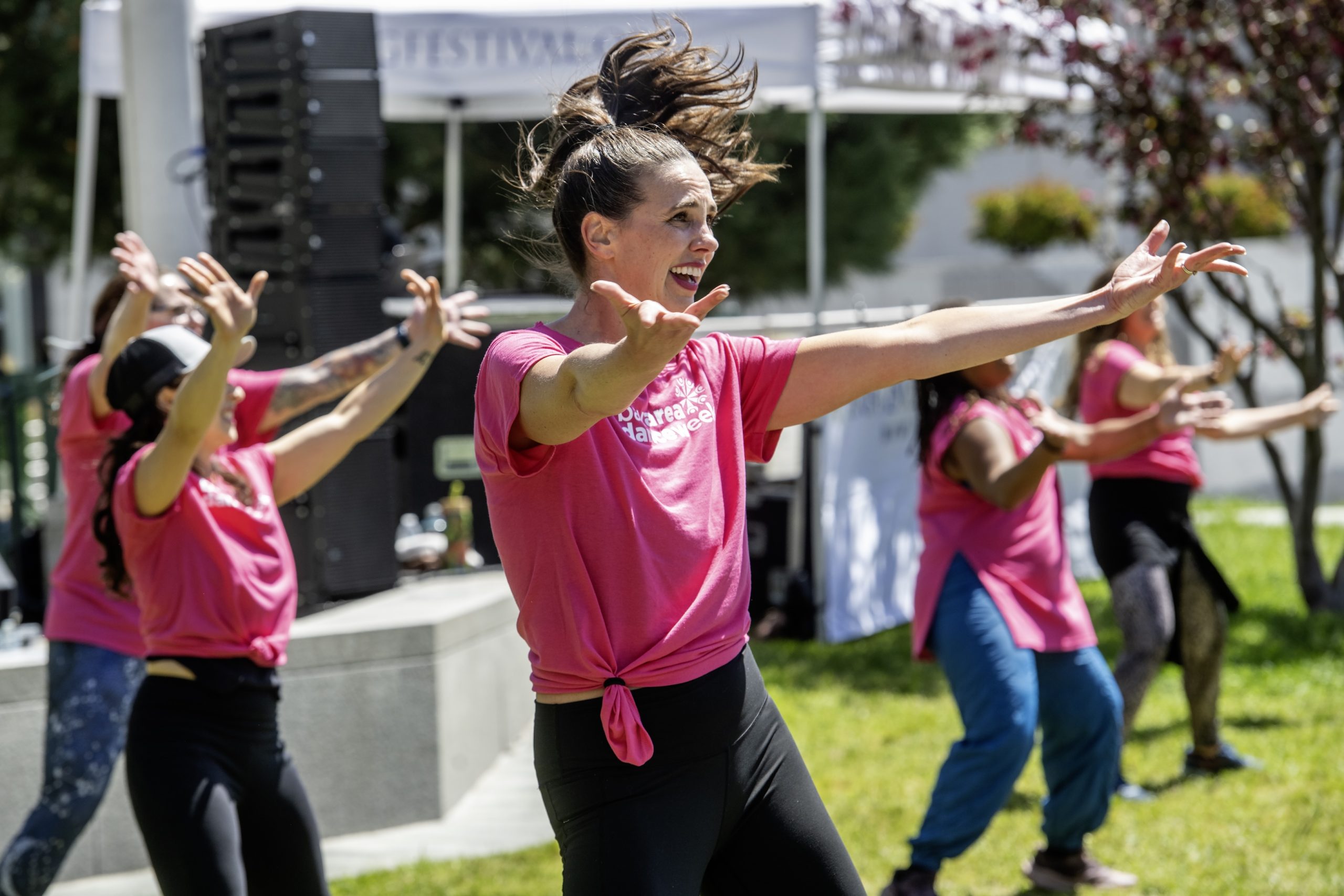 A group of women wearing pink shirts performs a dance routine outdoors on a sunny day.