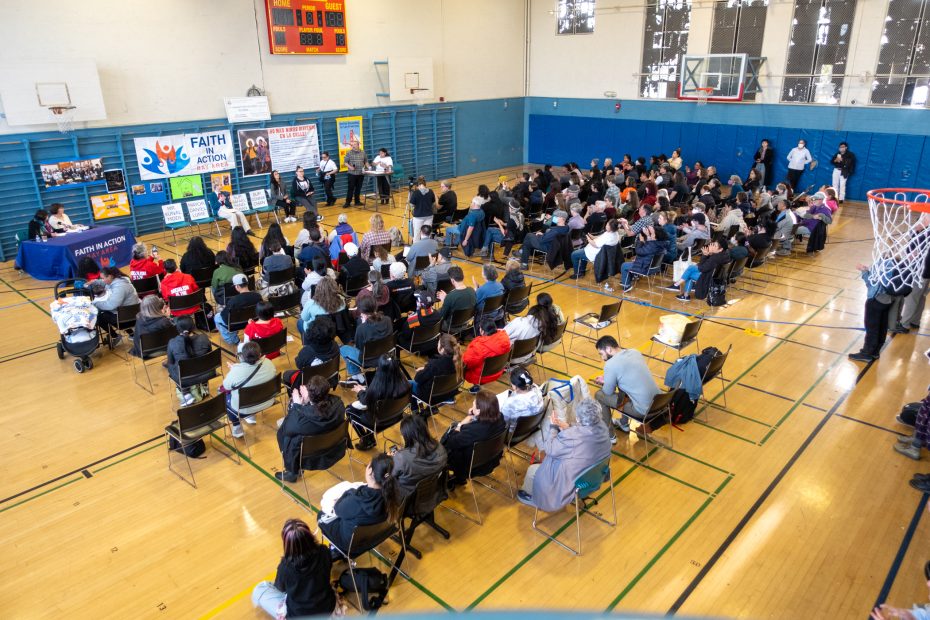 A large group of people seated in a gymnasium listens to a presentation. A banner on the stage reads "Faith in Action." Chairs are arranged in rows facing the speakers at the front.