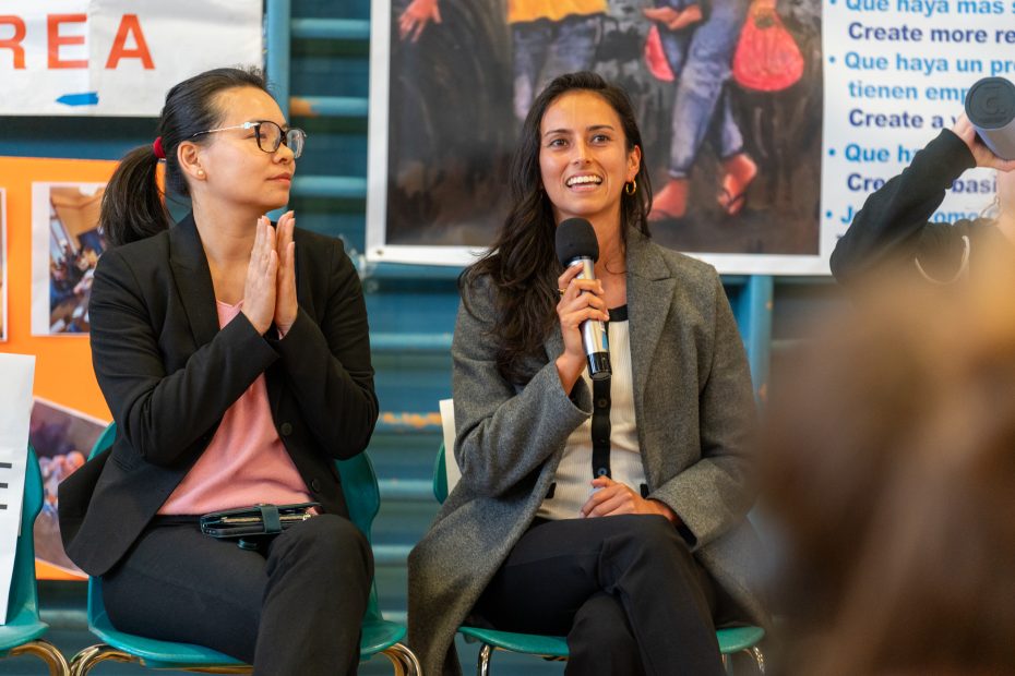 Two women sit on chairs; one holds a microphone and speaks, while the other claps. They are dressed in business attire, with posters visible in the background.