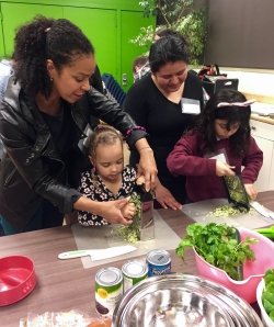 Two adults assist two children in grating vegetables at a kitchen counter. Cans, herbs, and a metal bowl are on the table.
