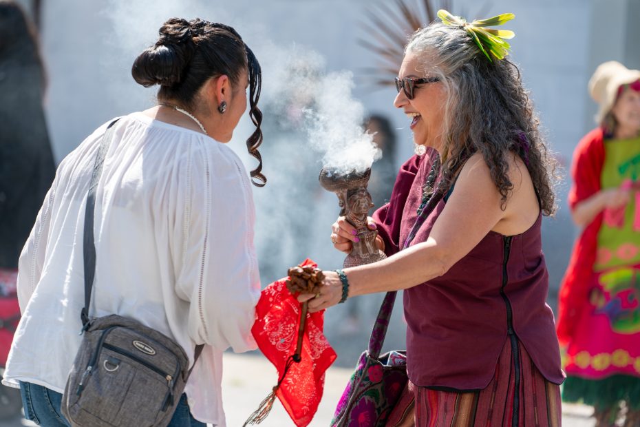 Two women participate in a cultural ritual involving smoke. One holds a smoking vessel, while the other leans in. They are surrounded by people and wear traditional attire.