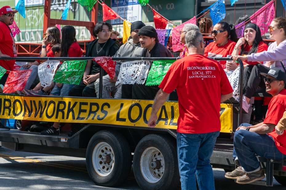 People wearing red shirts with "Ironworkers Local 377" text are seated on a parade float decorated with colorful flags.