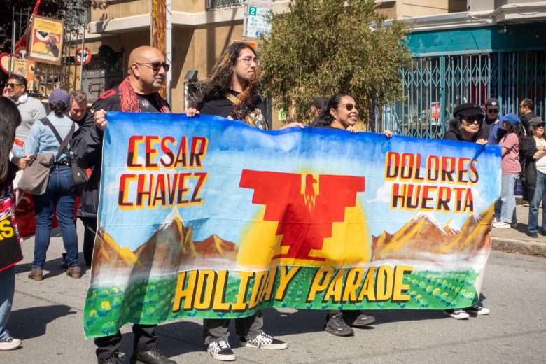 People hold a colorful banner with "Cesar Chavez," "Dolores Huerta," and "Holiday Parade" written on it during a street parade.