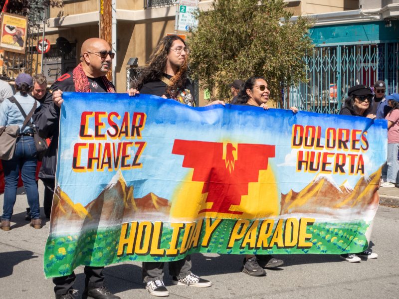 People hold a colorful banner with "Cesar Chavez," "Dolores Huerta," and "Holiday Parade" written on it during a street parade.
