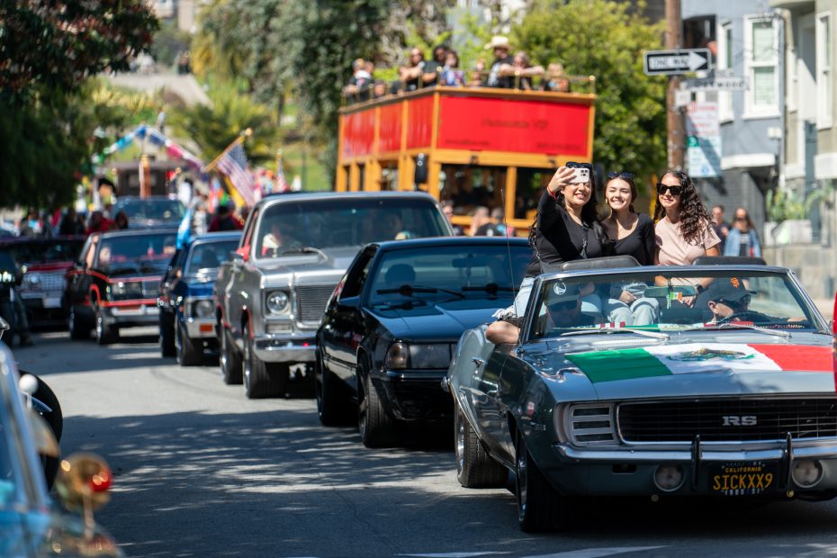 A parade of classic cars moves along a street. A woman takes a selfie in a convertible adorned with a Mexican flag while others ride and watch. A double-decker bus follows in the background.