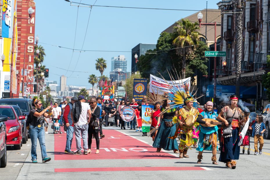 A cultural parade proceeds down a city street, featuring traditionally dressed individuals with feathered headdresses and drums, accompanied by onlookers and various banners.